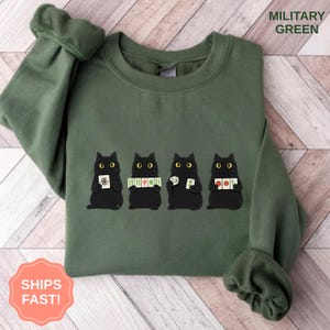May include: Military green sweatshirt featuring a graphic of four black cats holding game tiles. The text "MILITARY GREEN" is printed at the top right. The words "SHIPS FAST!" are printed on a pink circle at the bottom left.