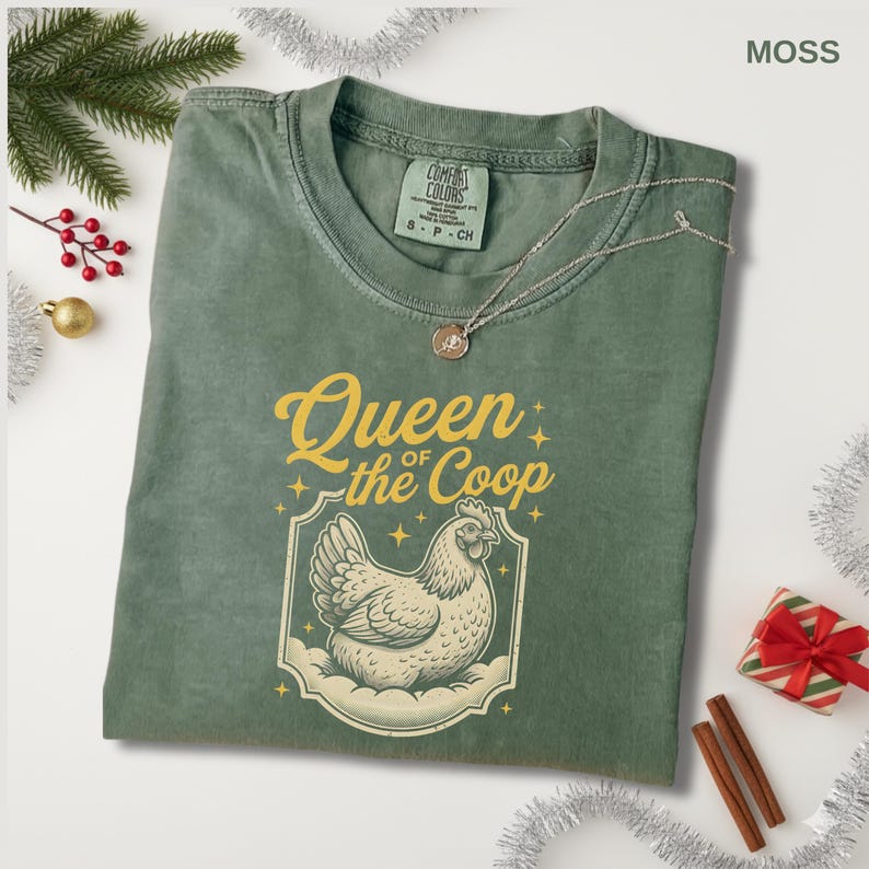 May include: Moss green t-shirt with the text "Queen of the Coop" and a graphic of a hen. The shirt has a crew neck and short sleeves. A silver necklace with a pendant is on the shirt.