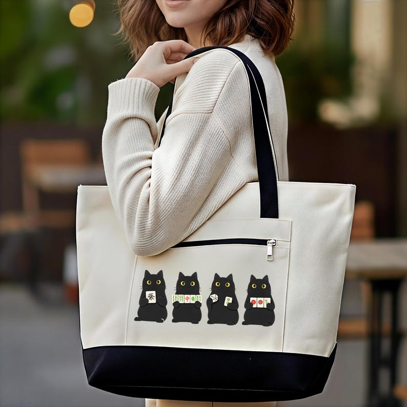 May include: Cream-colored tote bag with a black base and handles. The bag features a row of four black cat illustrations, each holding different items. A zippered pocket is visible on the front.