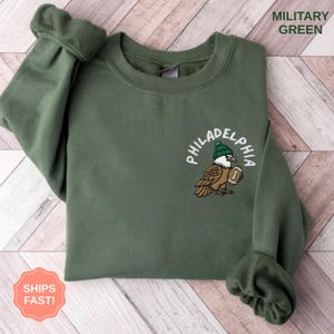 Peut inclure: Sweat-shirt vert militaire avec broderie d'un aigle de Philadelphie. L'aigle porte un bonnet vert et tient un ballon de football. Le mot "PHILADELPHIA" est inscrit en arc de cercle au-dessus de l'aigle. Le texte "MILITARY GREEN" est en haut à droite.