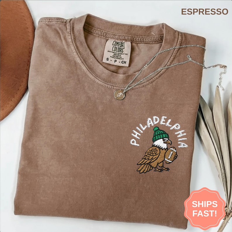 May include: Espresso-colored t-shirt with a crew neck. The shirt features an embroidered design of an eagle wearing a green beanie and holding a football, with the word "PHILADELPHIA" arched above. A silver necklace with a pendant is draped on the shirt.