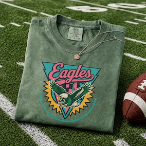 May include: A green Comfort Colors t-shirt with an Eagles logo, featuring a teal, pink, and yellow design of an eagle with outstretched wings. A silver necklace with a small pendant rests on the shirt, next to a brown football.