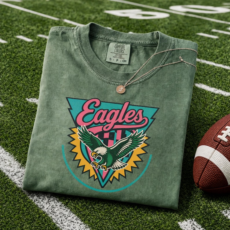 May include: A green Comfort Colors t-shirt with an Eagles logo, featuring a teal, pink, and yellow design of an eagle with outstretched wings. A silver necklace with a small pendant rests on the shirt, next to a brown football.