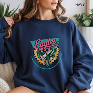 May include: Navy blue sweatshirt featuring a colorful Eagles graphic. The graphic includes the word "Eagles" in pink script, a teal triangle, and a green and white eagle with yellow accents. The sweatshirt is a casual style.