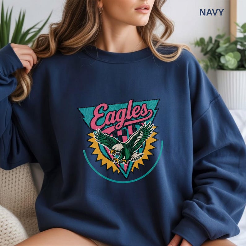 May include: Navy blue sweatshirt featuring a colorful Eagles graphic. The graphic includes the word "Eagles" in pink script, a teal triangle, and a green and white eagle with yellow accents. The sweatshirt is a casual style.