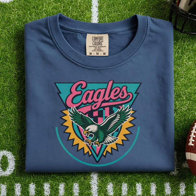 May include: A blue t-shirt with the word "Eagles" in pink script, a teal triangle, and a green and white eagle graphic. The shirt has a crew neck and a tag that says "Comfort Colors".