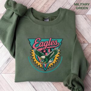 May include: A military green sweatshirt with the word "Eagles" in a retro design. The design includes a teal triangle, pink text, and a green eagle with yellow accents. The text "MILITARY GREEN" is printed in the upper right corner.