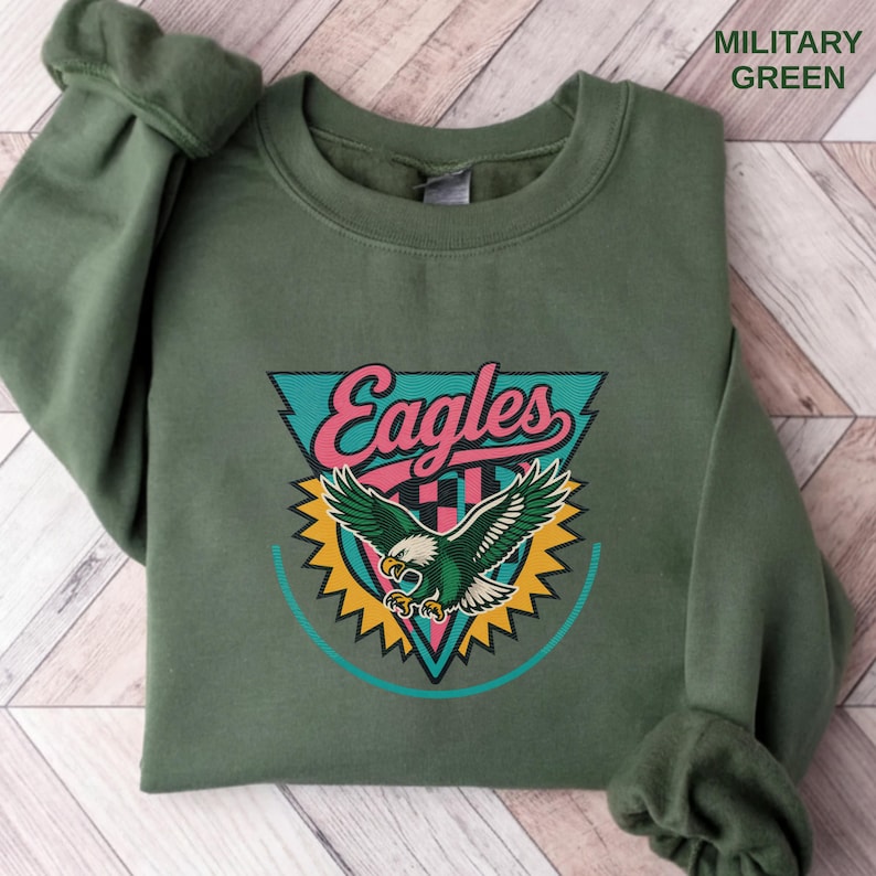 May include: A military green sweatshirt with the word "Eagles" in a retro design. The design includes a teal triangle, pink text, and a green eagle with yellow accents. The text "MILITARY GREEN" is printed in the upper right corner.