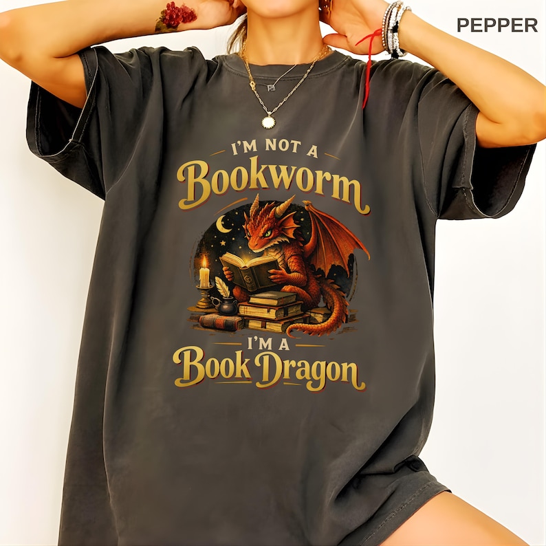 May include: Dark gray t-shirt with the text "I'm Not a Bookworm, I'm a Book Dragon." The graphic features a red dragon reading a book, with a candle and books. The design is in gold and the dragon has orange wings.