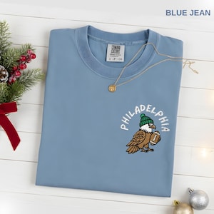 May include: A blue jean colored t-shirt with an embroidered Philadelphia eagle design. The eagle is wearing a green beanie and holding a football. A gold necklace with a pendant rests on the shirt.