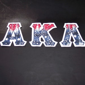 May include: Three decorative letters spelling "AKA" in a patriotic design. The letters are white with a blue background filled with white stars and red and white stripes. The letters are on a black surface.