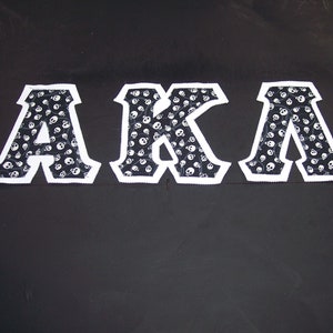 May include: White letters spelling "AKA" on a black background. The letters are made of fabric with a skull pattern.