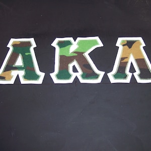 May include: A close-up of three large letters, "AKA", in a camouflage pattern with white outlines. The letters are sewn onto a black fabric background.