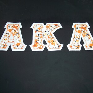 May include: White felt letters spelling "AKA" with orange and black splatter paint design.