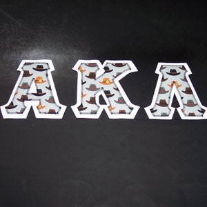 May include: White outlined letters spelling "AKA" with a gray background and a pattern of brown and orange cowboy hats.