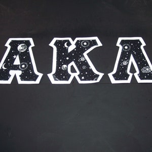 May include: Three embroidered patches spelling "AKA" in a varsity letter style. The letters are black with a white border and filled with a white and grey space-themed design featuring stars, planets, and moons. The background is black.
