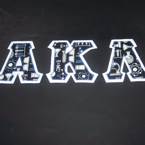 May include: White felt letters spelling "AKA" with a blue and white science themed pattern. The letters are on a black background.