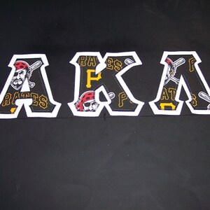 May include: White letters spelling "AKA" with a black background. Each letter is made of fabric with a pattern of a cartoon pirate holding a baseball bat and the words "Pirates" and "P" repeated.