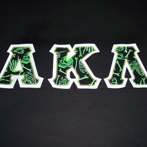 May include: Black fabric with white outlined letters spelling "AKA" in green. The letters are filled with a pattern of green skeletons and bones.
