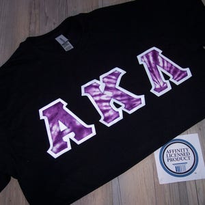 May include: Black t-shirt with a purple and white tie-dye design of the letters "AKA". The shirt has a white border around the letters. The shirt also has a small white tag with the text "Affinity Licensed Product".