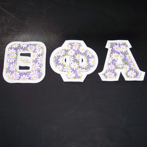 May include: Three fabric letters: a B, a Greek letter Phi, and an A. Each letter is filled with a pattern of small white daisies on a purple background. The letters have a white border and are set against a black background.