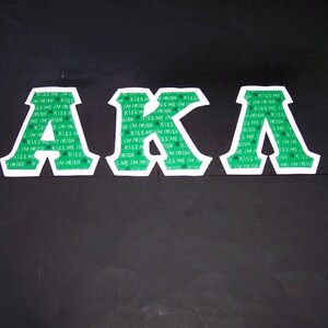 May include: Three white-outlined green letters spelling "AKA" on a black background. The green sections are patterned with the words "Kiss Me I'm Irish". The letters are likely patches or appliques.
