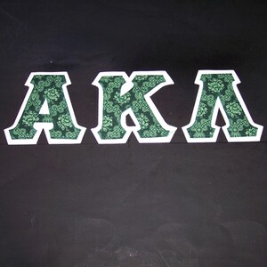 May include: The letters "AKA" are displayed against a black background. Each letter is white-bordered and filled with a green floral pattern. The letters are evenly spaced and centered.