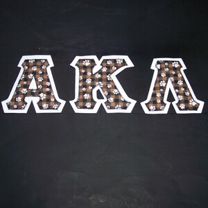 May include: The letters "AKA" are displayed against a black background. Each letter is white with a brown and black plaid pattern and white paw prints. The letters are outlined in white.