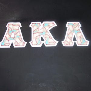 May include: Three white-bordered patches spelling "AKA". Each letter is filled with a baseball design, featuring red stitching and light blue accents. The patches are set against a dark background.