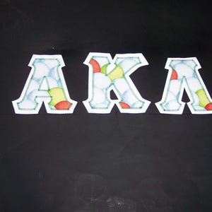 May include: White, stitched letters spelling "AKA" on a black background. The letters are filled with a pattern of white, blue, red, and yellow shapes. The letters have a white border with a green stitch.