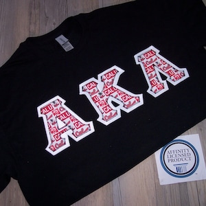 May include: Black t-shirt with the letters "AKA" in a white and red design. The letters are filled with a repeating pattern of red and white text. A white sticker with blue text is in the lower right corner.
