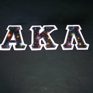 May include: Three embroidered patches spelling "AKA" in white-outlined, block letters. The letters are filled with a black background and colorful firework-like designs. The patches are displayed on a black surface.