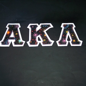 May include: Black and white letters spelling "AKA" with a colorful firework pattern inside each letter.