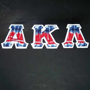 May include: Three fabric patches spelling "AKA". Each letter is white-bordered with a tie-dye pattern of red and blue. The patches are on a black background.