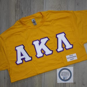 May include: A goldenrod yellow short-sleeve t-shirt featuring the Greek letters AKA in white with purple outlines. A small white rectangular label on the shirt reads GOLD (GOLDENROD) SHIRT, and a circular sticker reads AFFINITY LICENSED PRODUCT.