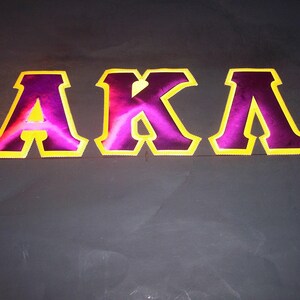 May include: The letters "AKA" in a bold, stylized font. The letters are a vibrant purple with a yellow outline, set against a dark gray background. The design is eye-catching and graphic.