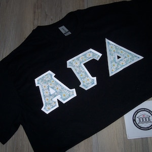 May include: Black t-shirt with a white floral design that spells out "AGA".