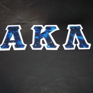 May include: Three embroidered patches spelling "AKA". Each letter is a white-bordered, navy blue shape filled with a starry, galaxy-like pattern. The patches are displayed on a black background.