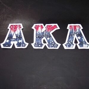 May include: Three decorative letters spelling "AKA" with a patriotic design. The letters are white-edged with a red, white, and blue pattern featuring stars and stripes. The letters are displayed against a dark background.