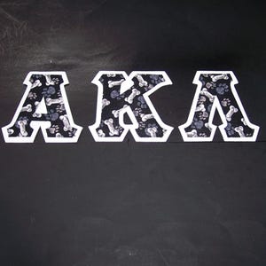 May include: The letters "AKA" are displayed in a bold, blocky font. Each letter is white with a black and white patterned interior featuring paw prints and bones. The background is a solid black.