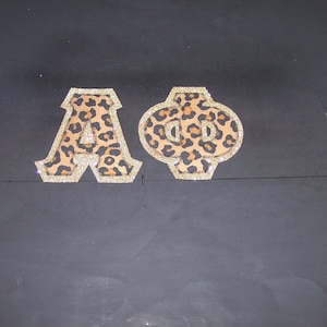 May include: Two decorative patches featuring the letters "A" and "Φ". The letters have a leopard print design with a gold glitter border. The patches are set against a dark background.