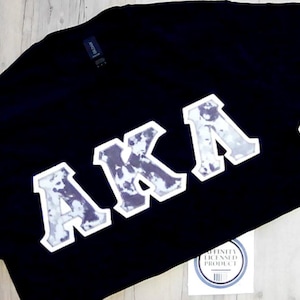 May include: Black t-shirt with the letters "AKA" in a white and gray patterned design. The shirt has a tag that says "Affinity Licensed Product".