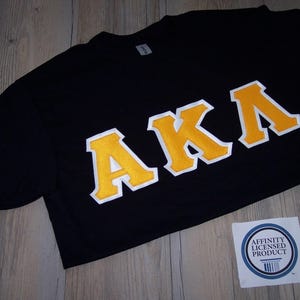 May include: Black t-shirt with the letters "AKA" in yellow with white trim. The shirt is lying on a wooden surface. A small sticker with the text "Affinity Licensed Product" is visible.