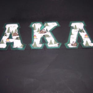 May include: Three fabric letters spelling "AKA" with a camping theme. Each letter is white with a pattern of cabins, trees, and campfires, outlined in green. The letters are displayed against a dark background.