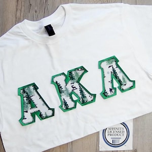 May include: White t-shirt with the letters "AKA" in a green and white forest design. The letters are outlined in green and have a textured appearance. A small tag with text is visible in the bottom right corner.