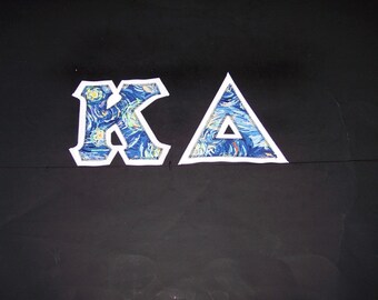 May include: Two Greek letters, Kappa and Delta, are appliqued onto a solid black fabric. The letters feature a blue, yellow, and white swirling pattern reminiscent of a starry night sky, each outlined with a thick white border and stitching.
