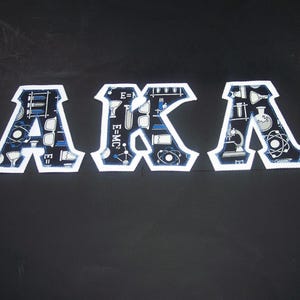 May include: Three white-bordered patches spelling "AKA". Each letter is filled with a blue and white science-themed pattern, including beakers, atoms, and the equation E=MC². The patches are on a dark background.