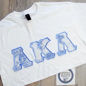 May include: White t-shirt with the letters "AKA" in a light blue, cloud-like design. The letters have a bold, outlined appearance. A small tag with text and a barcode is visible at the bottom.