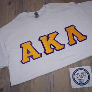 May include: White t-shirt with the letters "AKA" in gold and purple. The shirt is made of a soft material and is likely for casual wear. A small sticker with the text "Affinity Licensed Product" is in the bottom right corner.