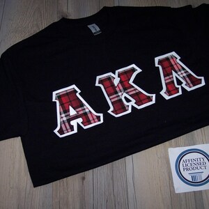 May include: Black t-shirt with the letters "AKA" in a white outline with a red and black plaid pattern. The shirt has a crew neck and short sleeves. A small white sticker with blue text is in the bottom right corner.